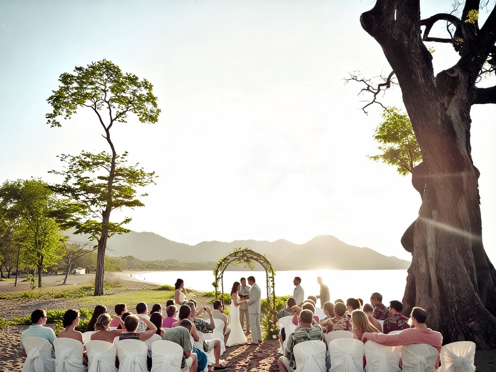 A destination wedding ceremony held outdoors on a beach, with guests seated and surrounded by scenic trees and majestic mountains; also consider the overall destination wedding cost.