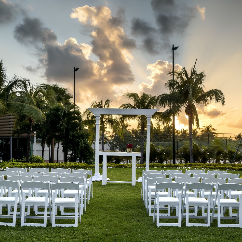White chairs set up for a destination wedding at sunset, framed by palm trees in the background.