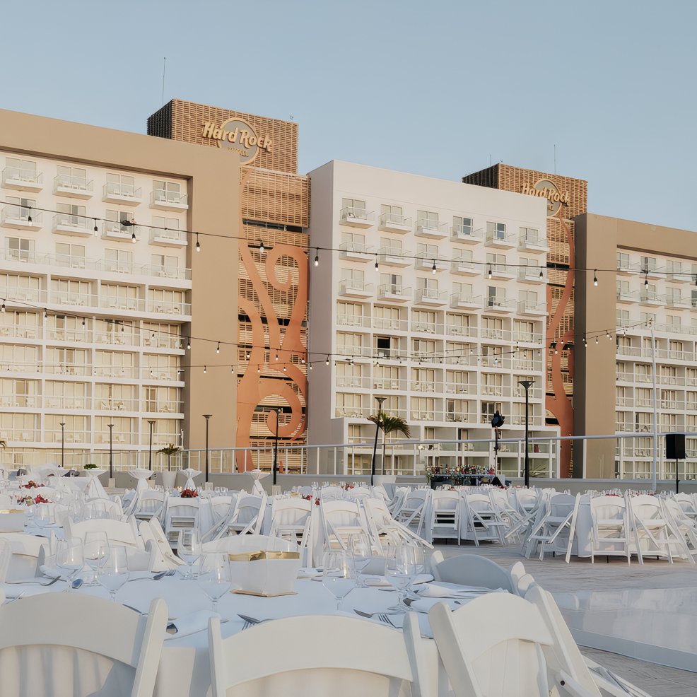 White tables and chairs arranged outdoors for a destination wedding in front of a multi-story hotel.