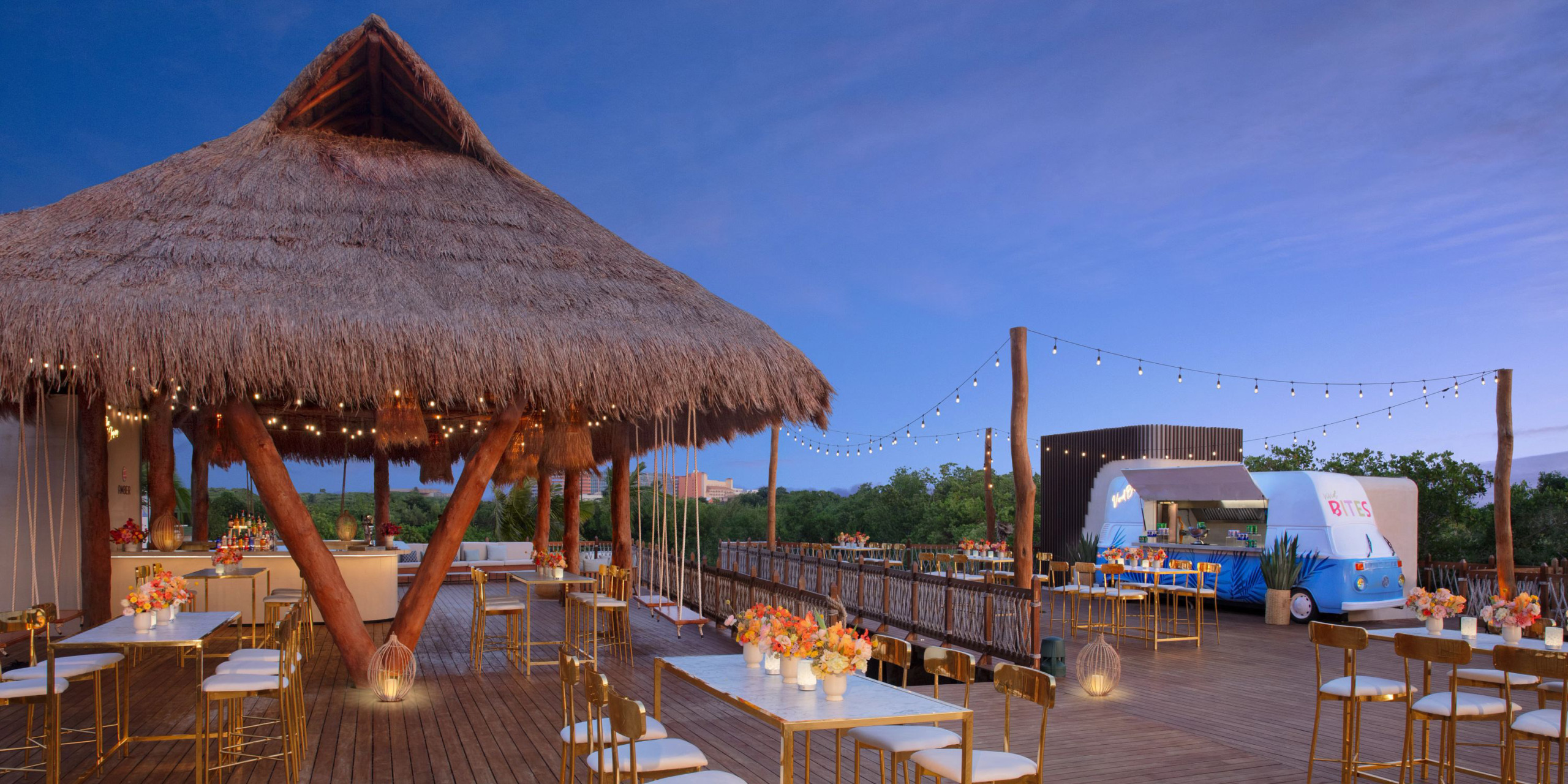 Outdoor dining area at the Hyatt Vivid Grand Island, featuring a thatched roof structure, string lights, and tables adorned with flowers. A food truck can be seen in the background, amidst lush greenery under a clear blue sky.