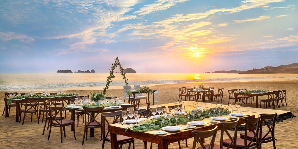 At Sunscape Dominicus La Romana, an outdoor beach wedding setup features tables, chairs, and plates arranged on the sand. An arch adorned with greenery faces the ocean as the sun sets over the water in the background.