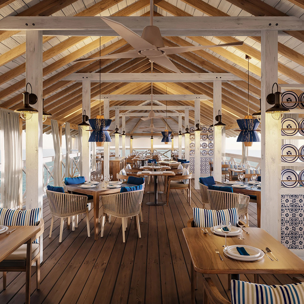 A cozy restaurant with wooden tables and striped cushions. The space features high ceilings with exposed beams, hanging lanterns, and large windows offering an ocean view.