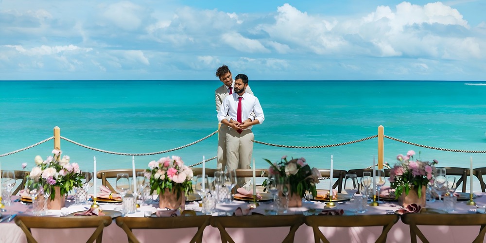 Two men stand by the ocean, behind a beautifully set table prepared for a destination wedding celebration.