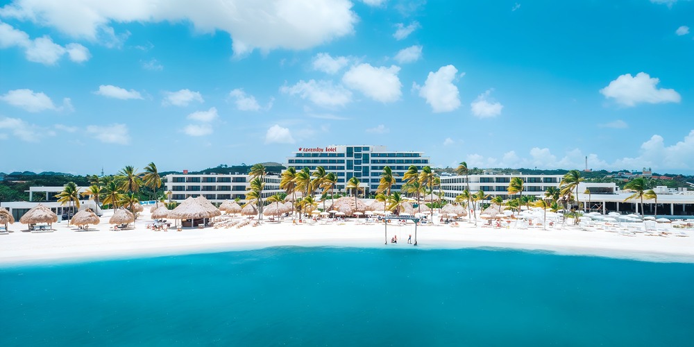Mangrove Beach Corendon Curacao is a multi-story beachfront hotel set among palm trees. In front of it lies white sand, thatched umbrellas, and the serene blue sea.