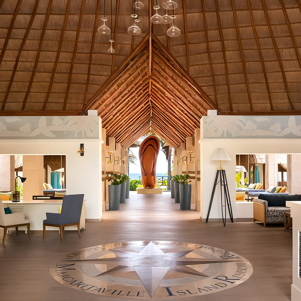Bright, open lobby with high wooden ceiling, modern furnishings, and a central surfboard display. A compass rose decorates the floor. Large windows show an ocean view.