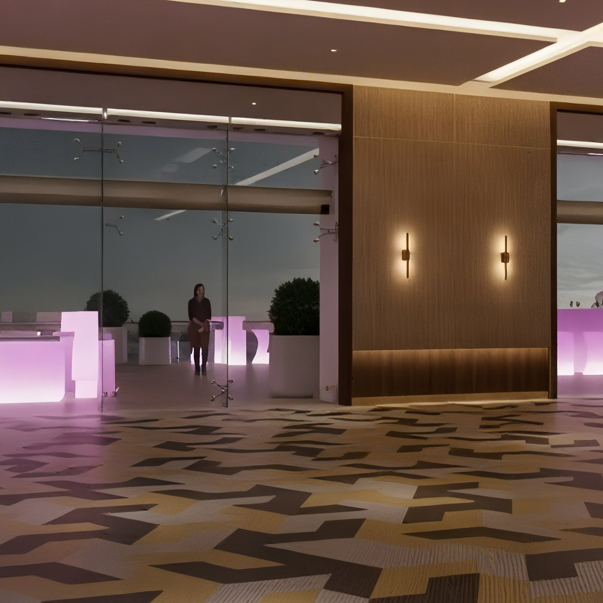 A modern lobby with geometric patterned flooring, glass walls, and illuminated counters. A person stands near a large window in the background.