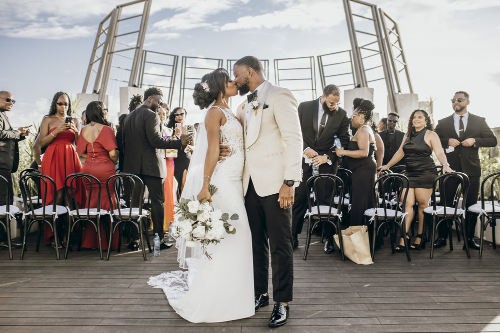 Carina and Marvin share a kiss on a deck at their destination wedding, surrounded by elegant guests.
