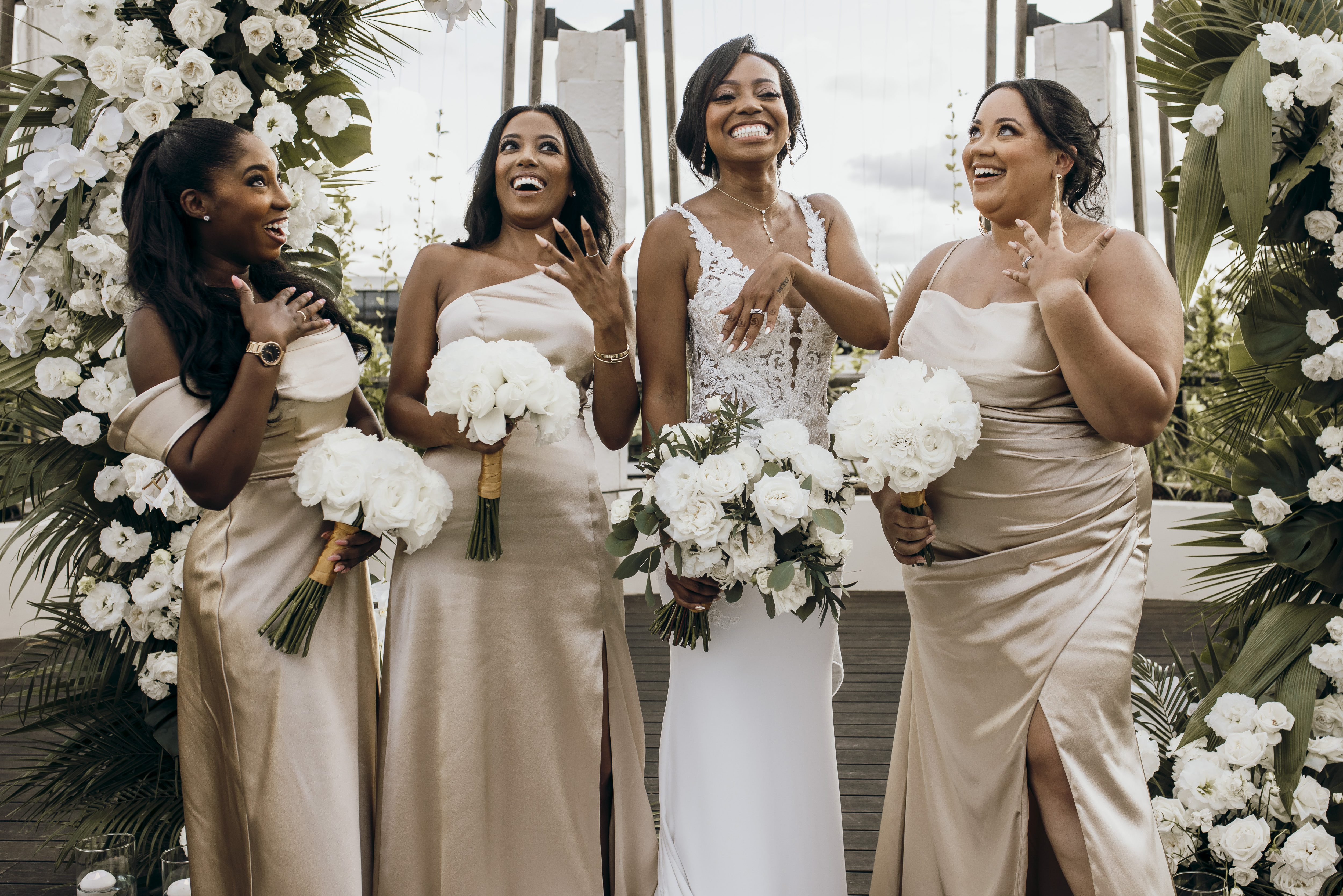 Four women in formal dresses show Carina and Marvin rings at a destination wedding, holding white bouquets.