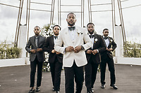 Five men in Carina and Marvin suits pose outdoors at a destination wedding with floral arrangements.