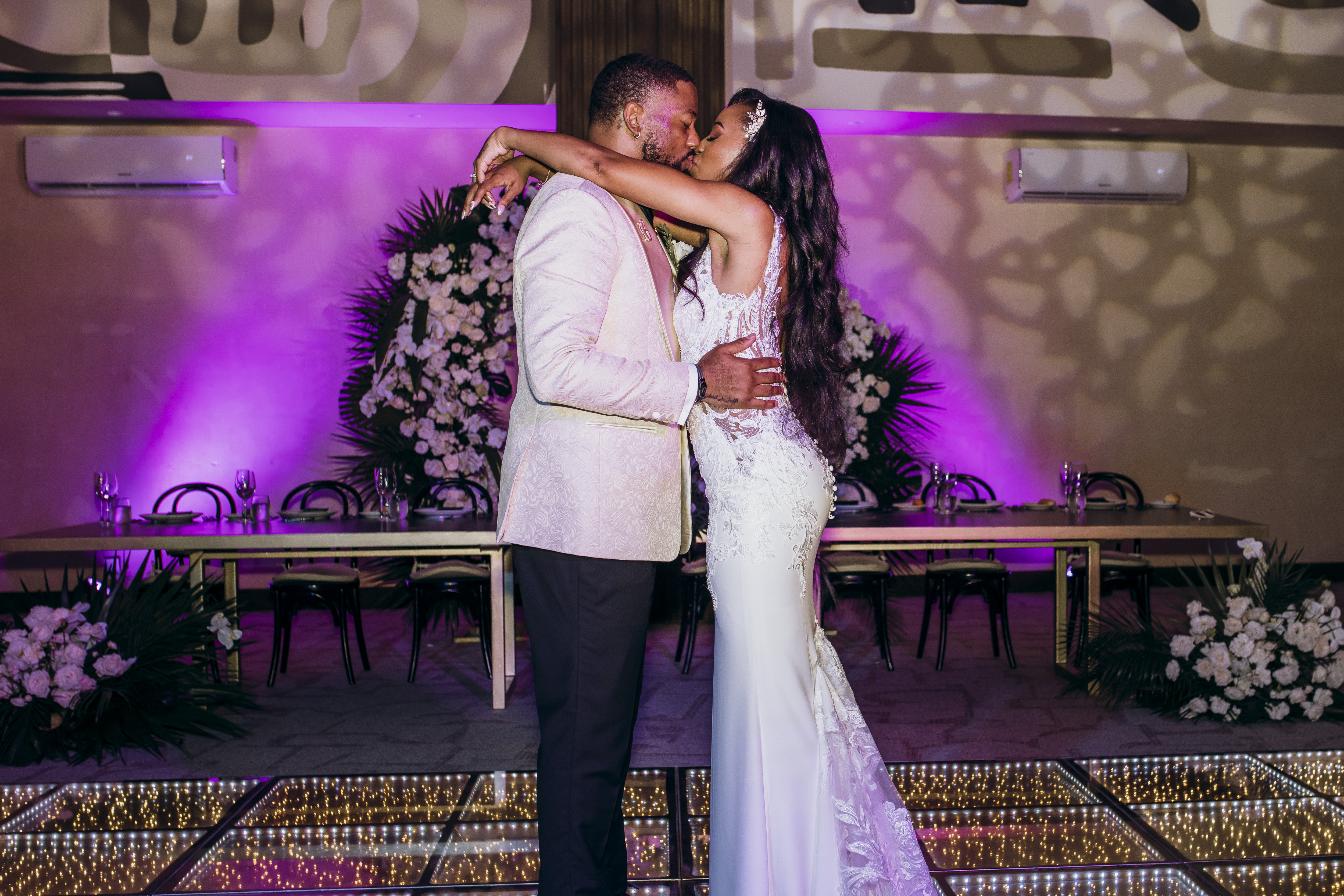 Carina and Marvin share a kiss at their destination wedding on a floral dance floor with purple lighting.