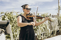 At a destination wedding, a woman in a black dress plays violin outdoors, surrounded by lush plants.