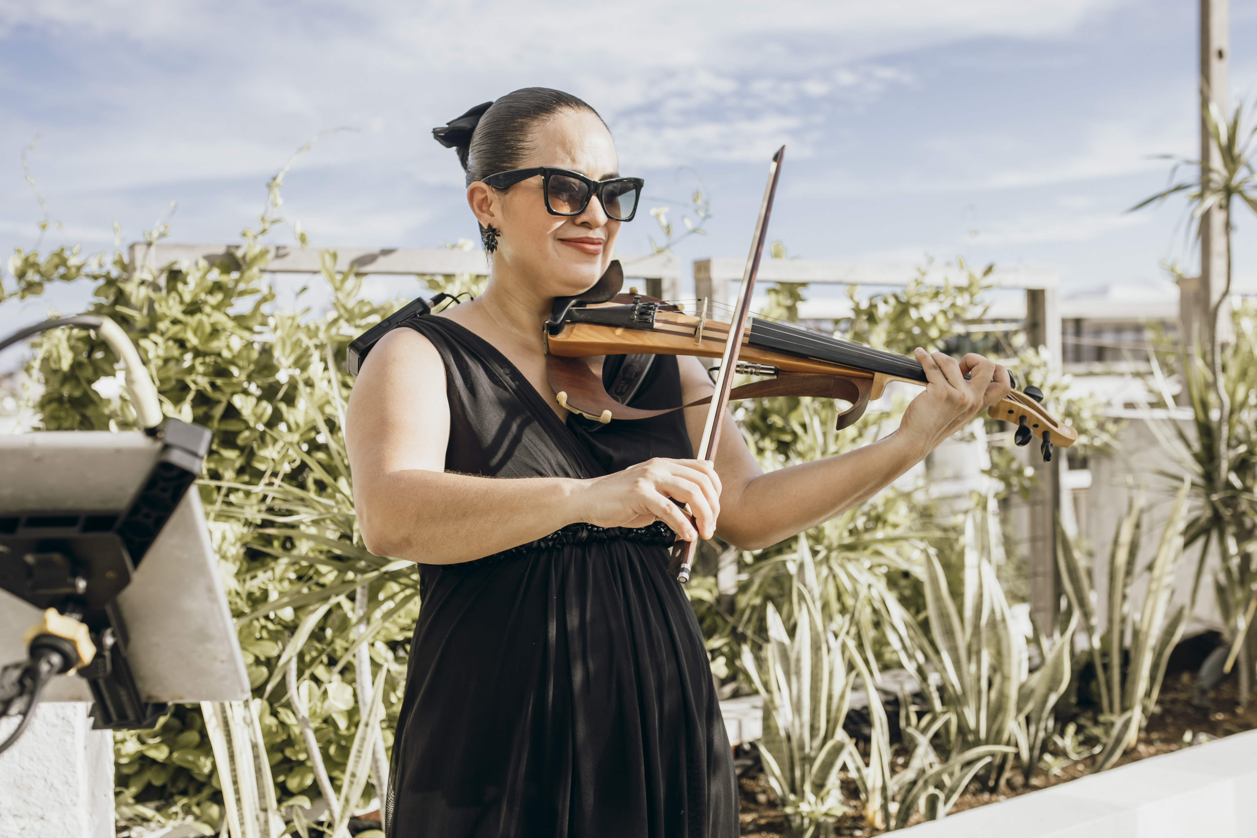 At a destination wedding, a woman in a black dress plays violin outdoors, surrounded by lush plants.