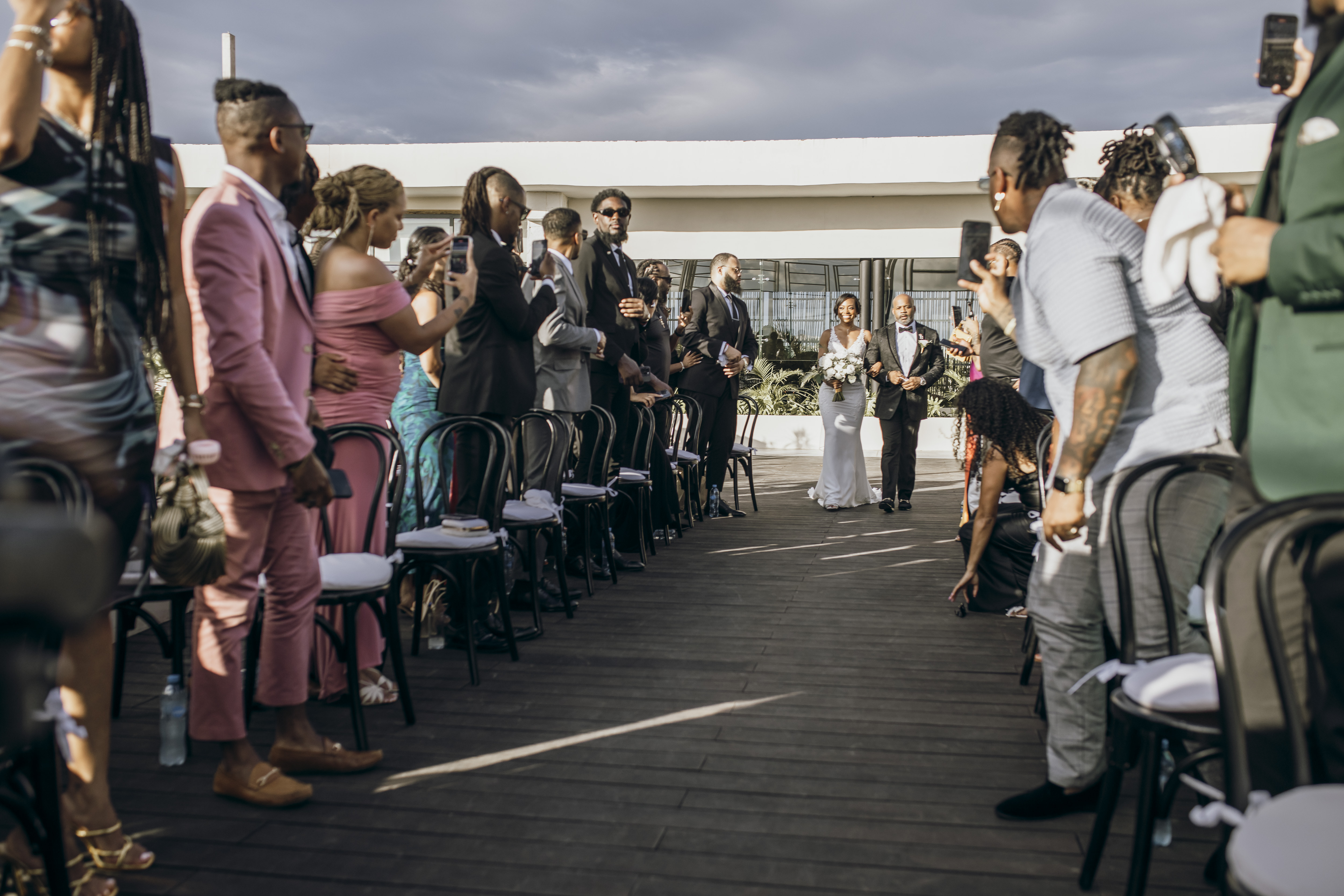 Carina and Marvin walk down the aisle at their destination wedding as guests stand and take photos.