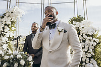 At a destination wedding, a man in a white tuxedo looks emotional, surrounded by lush white flowers.