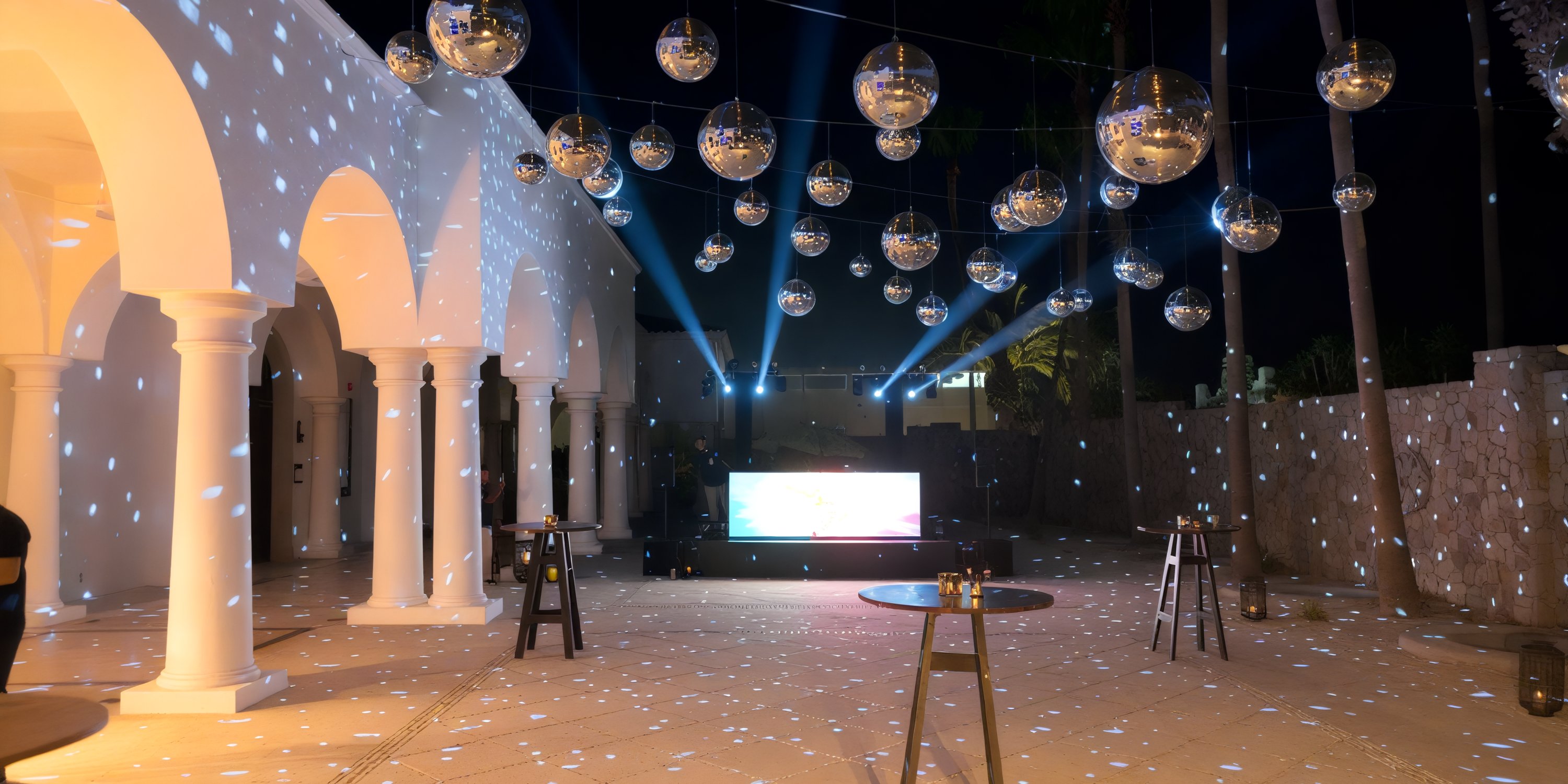 An enchanting outdoor event space for a destination wedding, featuring shimmering disco balls, elegant spotlights, beautifully arranged tables, and stunning arched columns all illuminated under the night sky.