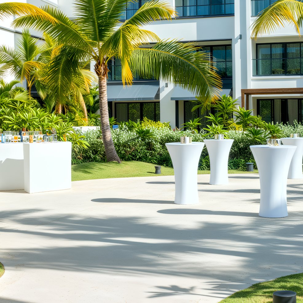 Outdoor cocktail tables and a stylish drink station are set up near a modern building with palm trees, creating an inviting atmosphere for a destination wedding.