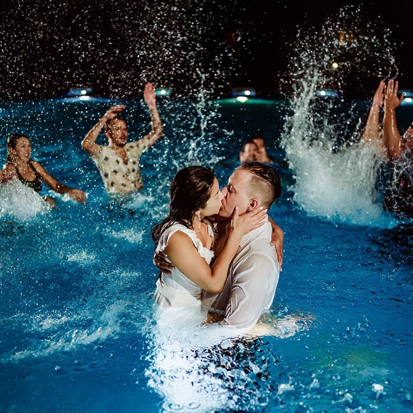 Clarissa and Jon share a kiss in the pool under the night sky, surrounded by friends splashing water during their destination wedding celebration.