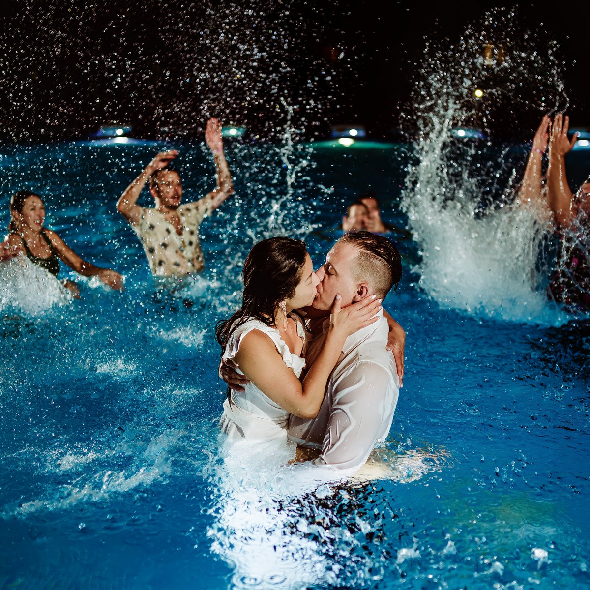 Clarissa and Jon share a kiss in the pool under the night sky, surrounded by friends splashing water during their destination wedding celebration.