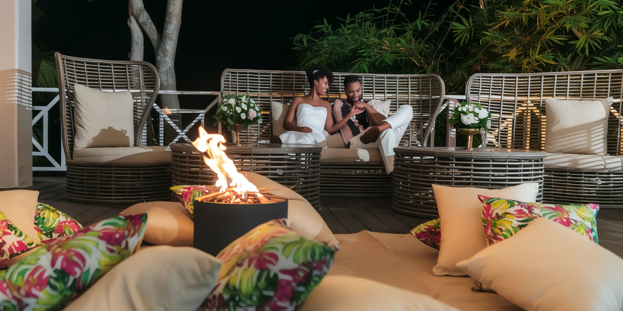 A couple relaxes on patio furniture by a fire pit, surrounded by plush cushions and lush plants, as they dreamily discuss plans for their upcoming destination wedding.