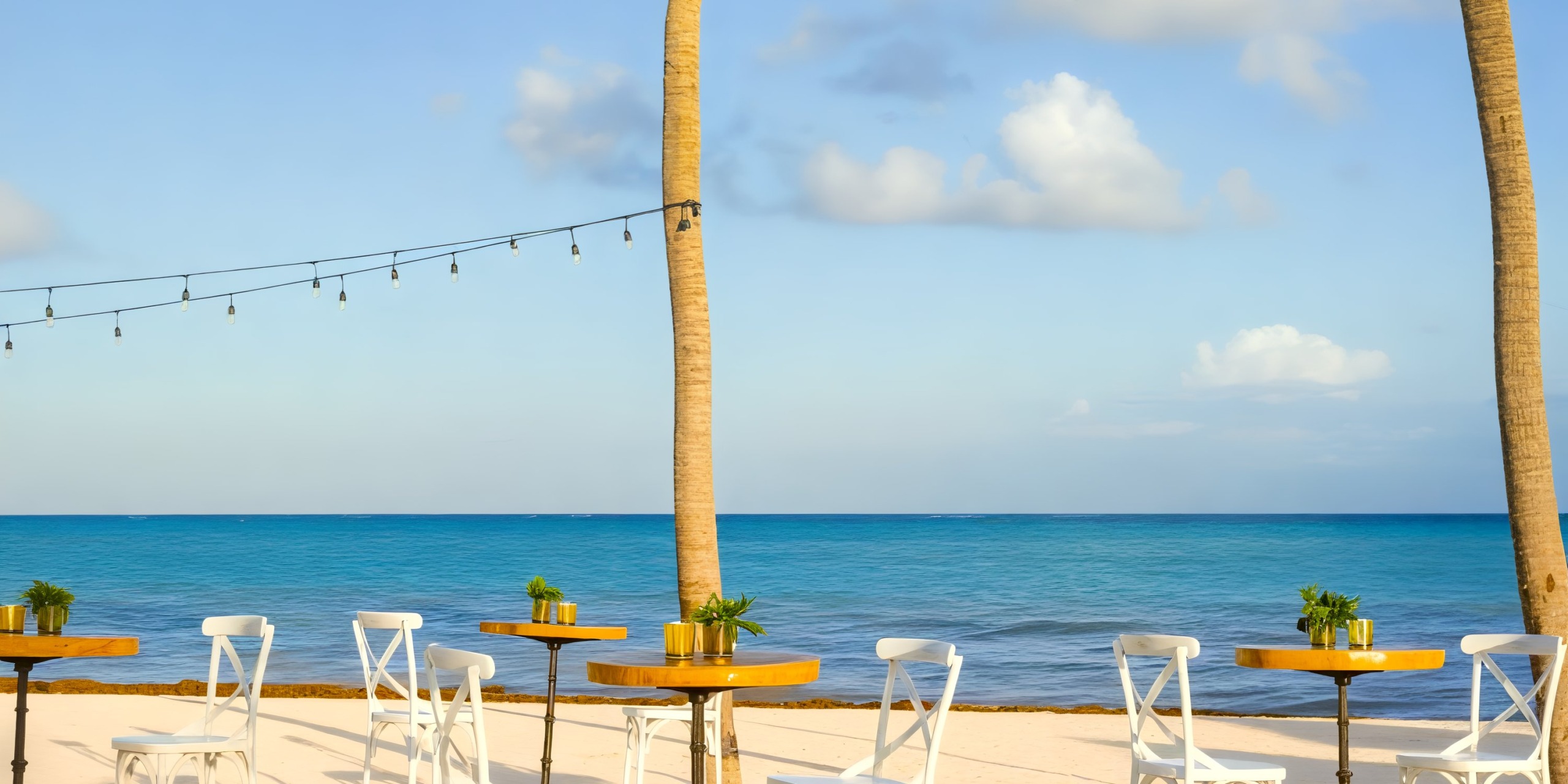 Experience a breathtaking destination wedding at Dreams Cap Cana Resort, featuring an oceanfront setup with elegant white chairs, round tables, and enchanting string lights.