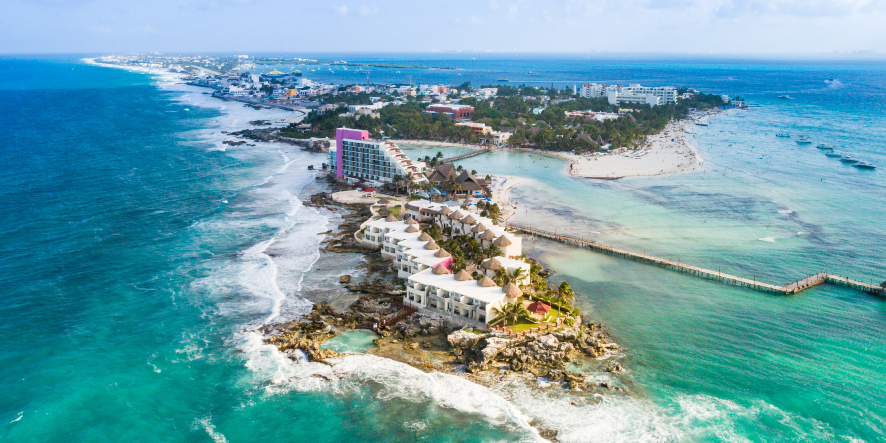 Aerial view of a coastal resort with buildings surrounded by turquoise sea and rocky shores, located on an island with visible beaches and a pier.