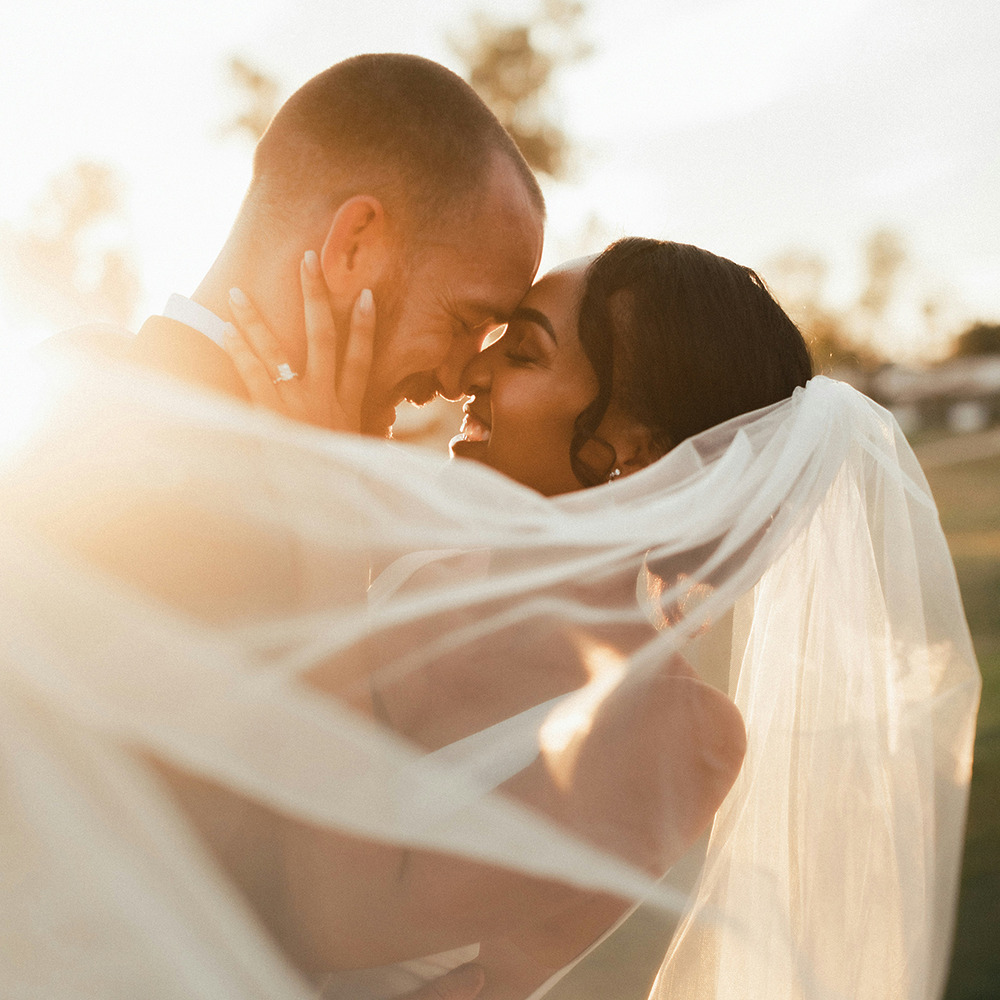 The radiant bride and groom share an embrace, their veil elegantly flowing in the sunlight, capturing the enchanting essence of a destination wedding.