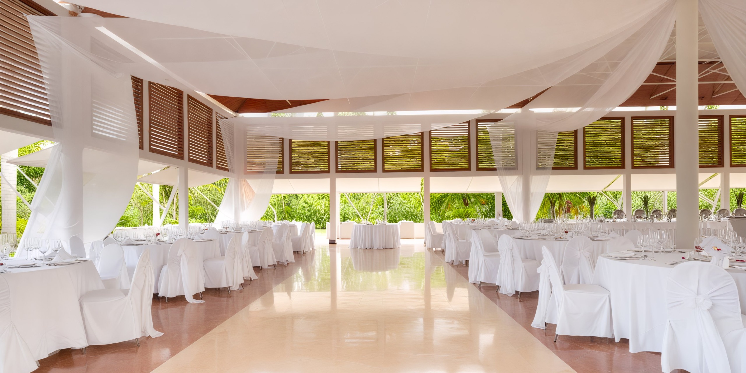 The interior of an elegant, empty banquet hall, adorned with white tables and sheer drapery, perfectly sets the scene for a stunning destination wedding.