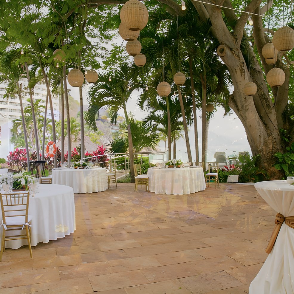 Chic wedding event space outdoors with round tables, gold chairs, and lanterns hanging from trees.