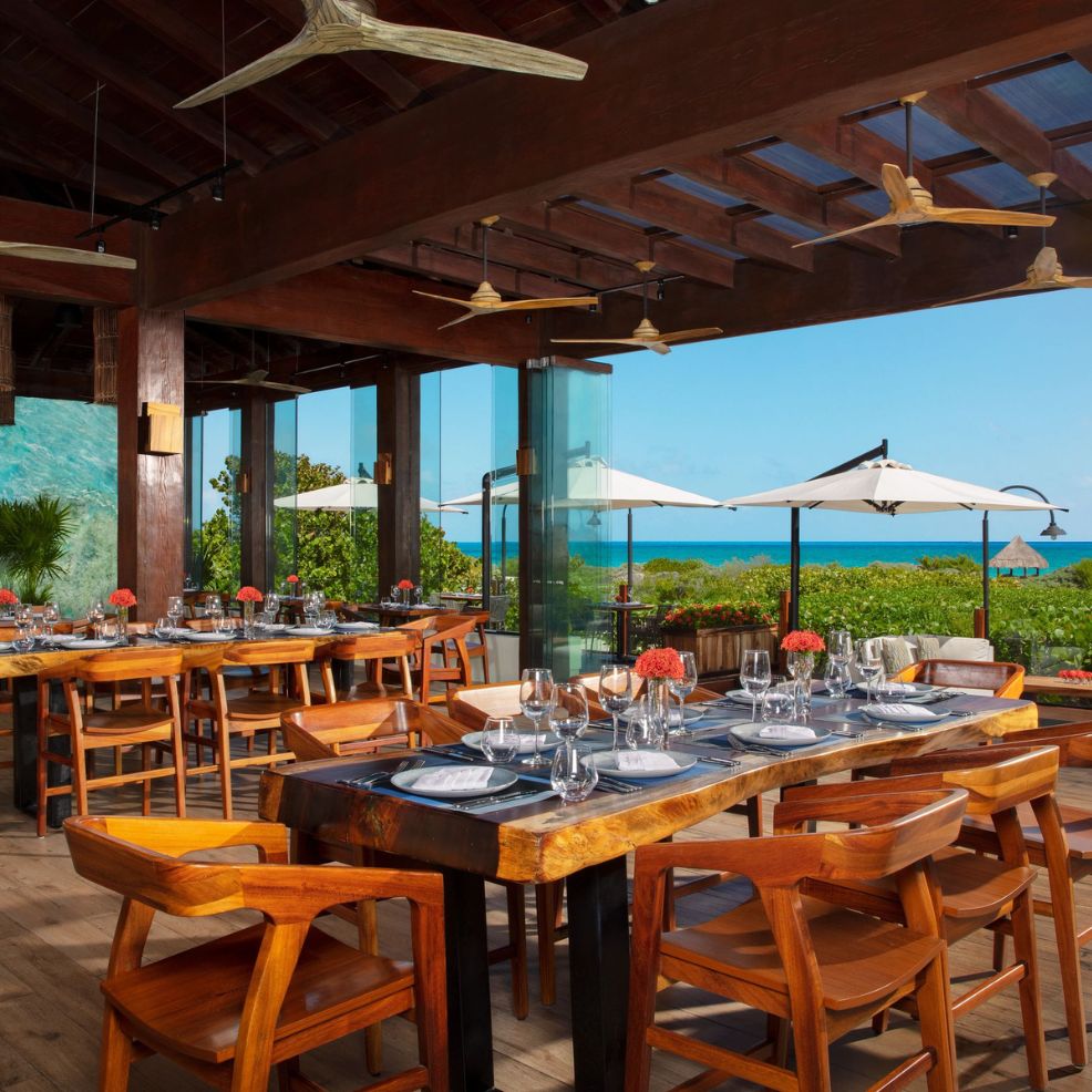 An open-air restaurant featuring wooden tables elegantly set for dining, offering a breathtaking ocean view through glass walls, makes for an ideal destination wedding venue.