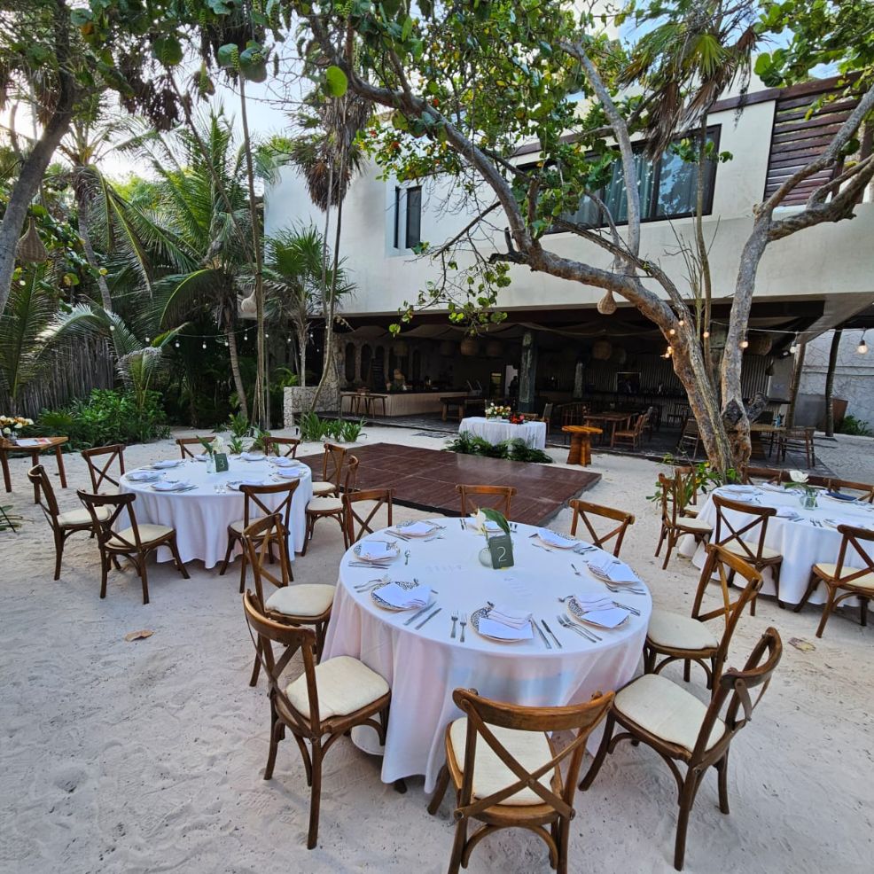 Round tables with white cloths arranged on sand for a destination wedding near a building and trees.