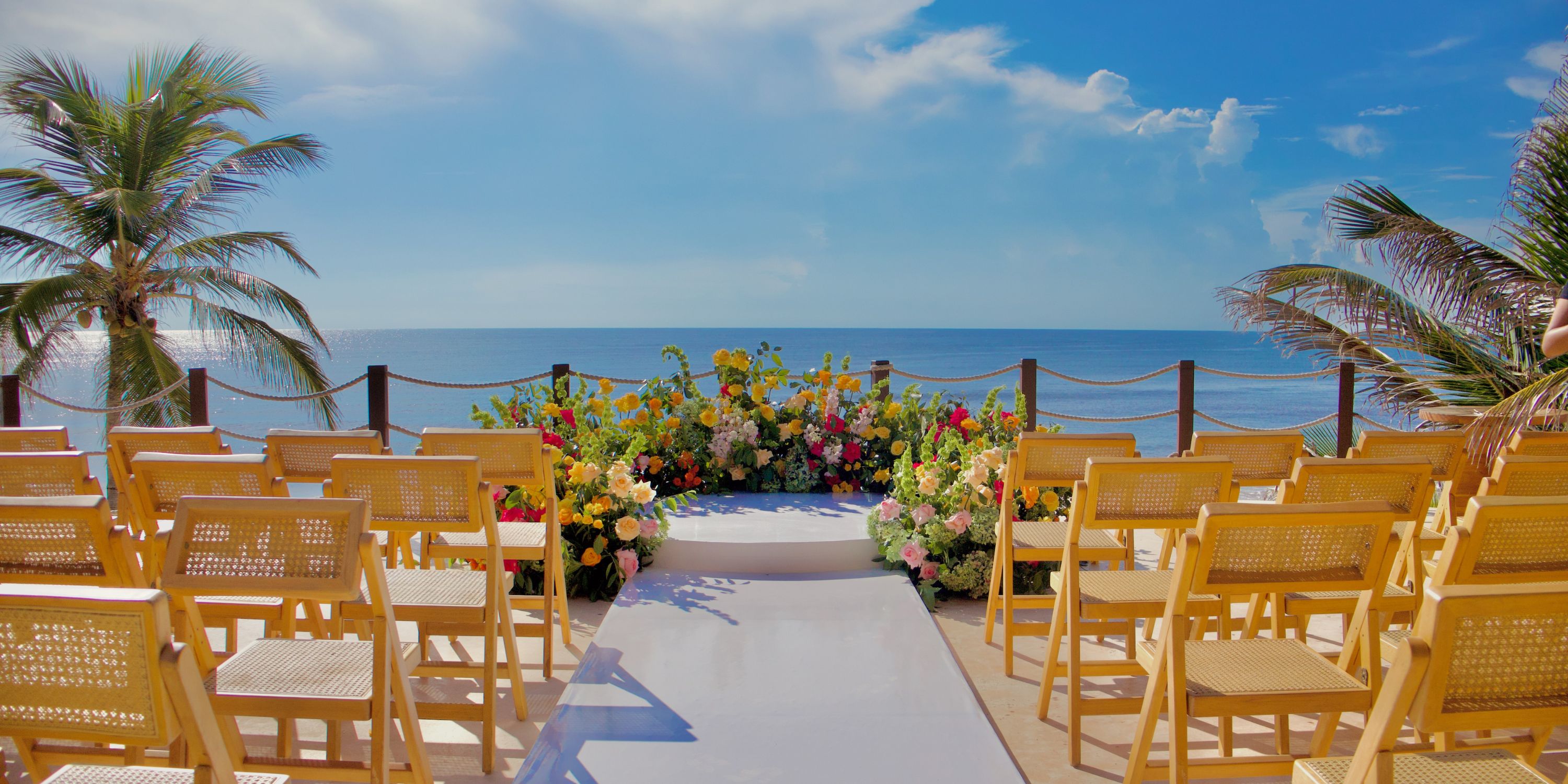 Rows of yellow chairs face a floral wedding altar at a destination wedding by the ocean under sunny skies.
