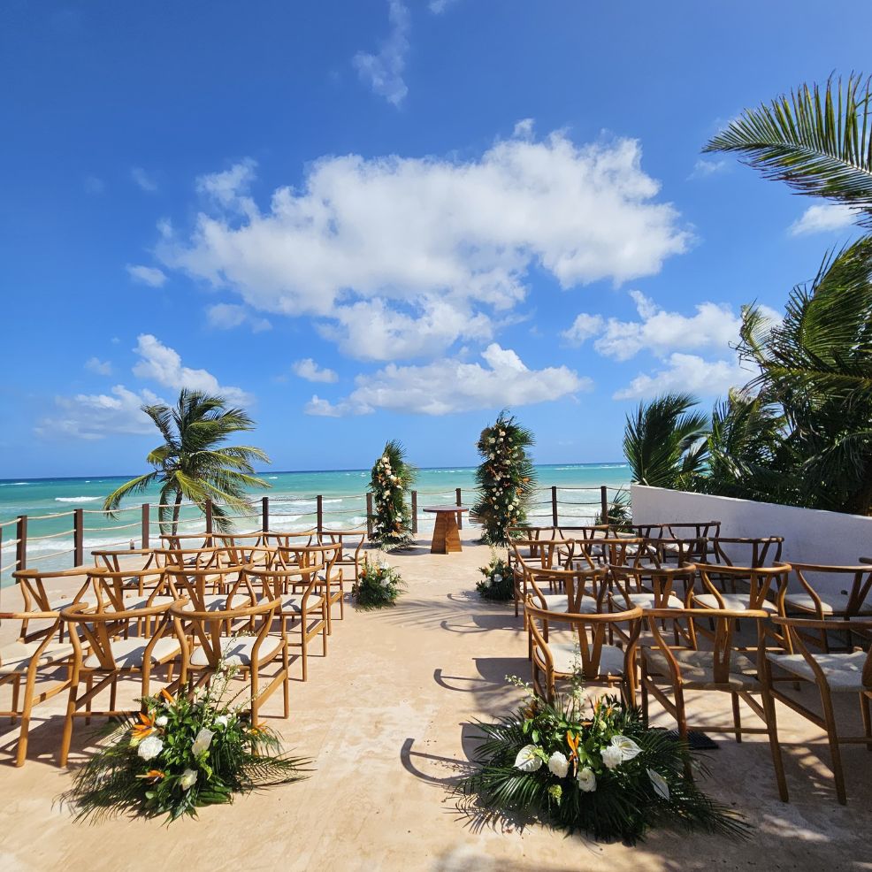 Destination wedding ceremony by the ocean, with wooden chairs and floral decor arranged on the beachfront.