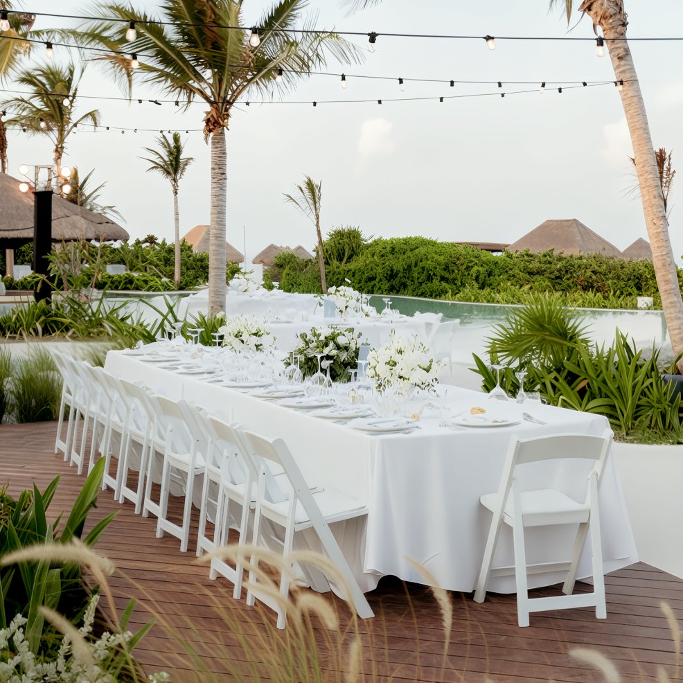 Long dining table elegantly set for a destination wedding, surrounded by palm trees and lush greenery.
