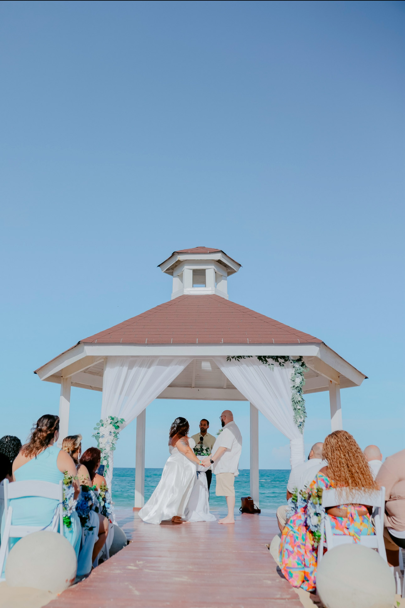 A couple is having their destination wedding under a gazebo by the ocean, with guests seated nearby to witness the ceremony.