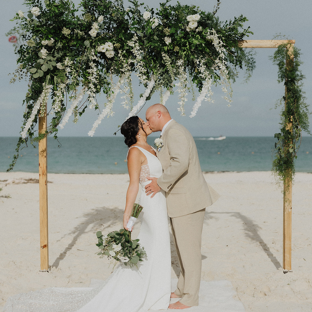 Chelsey and Zach share a kiss beneath a floral arch during their destination wedding on the beach.