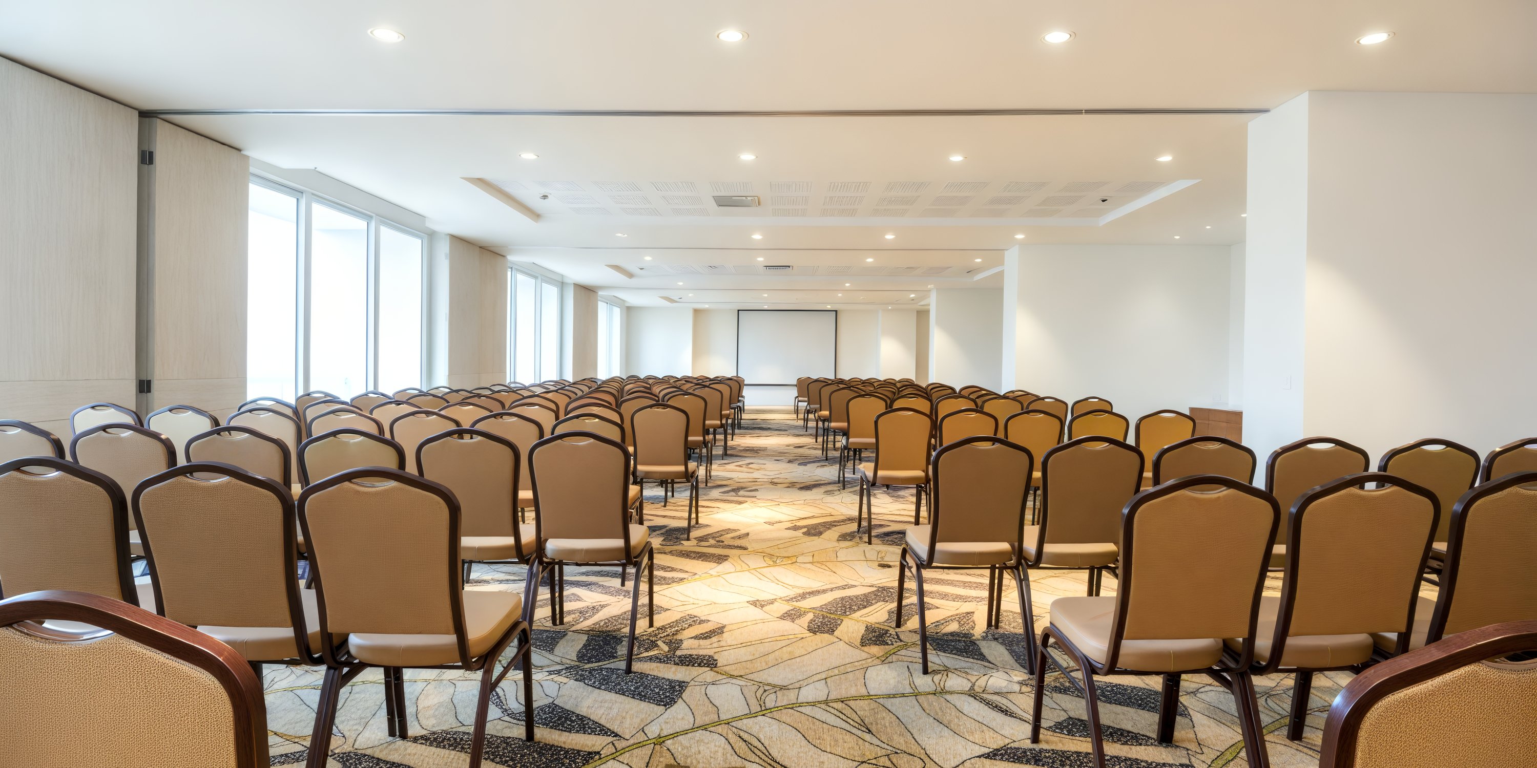 A vacant conference room adorned with rows of beige chairs, all oriented towards a screen, awaits transformation into the perfect venue for a memorable destination wedding.