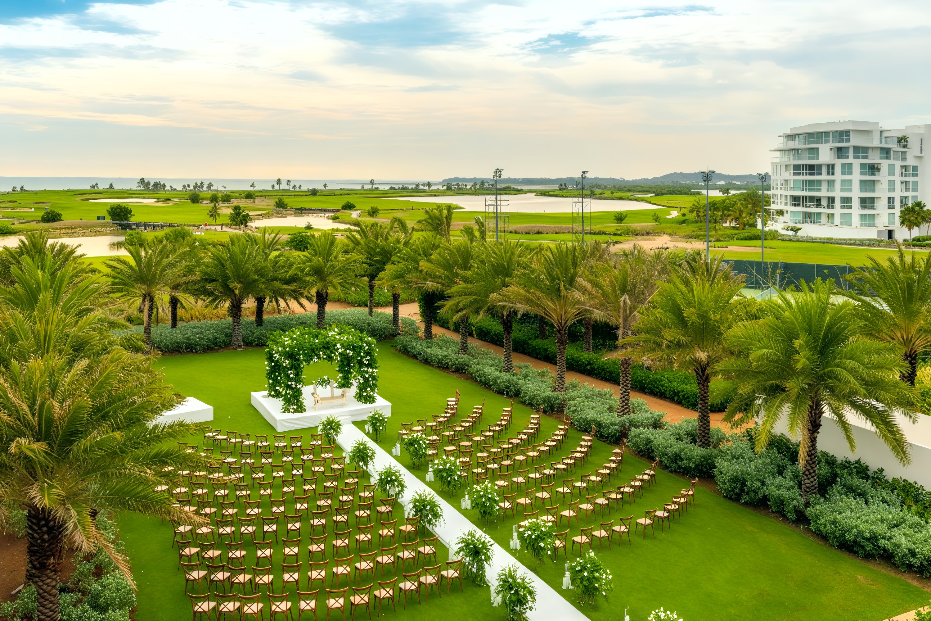 A picturesque destination wedding setup features an outdoor arrangement with chairs and an altar, elegantly surrounded by lush palm trees and vibrant greenery, all nestled near a charming building.