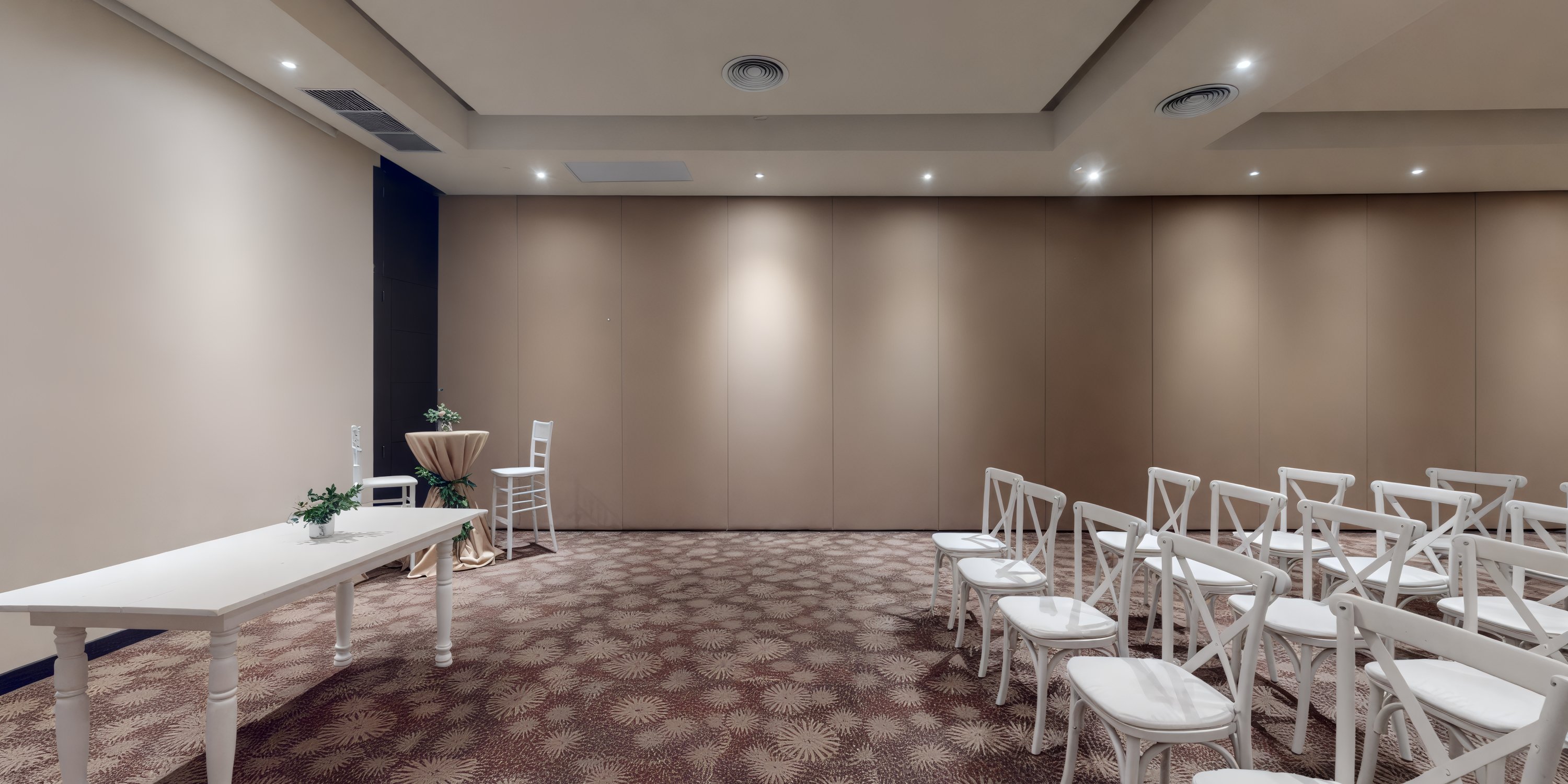 An unadorned space set for a wedding, featuring rows of white chairs and a table with one chair against soft beige walls.