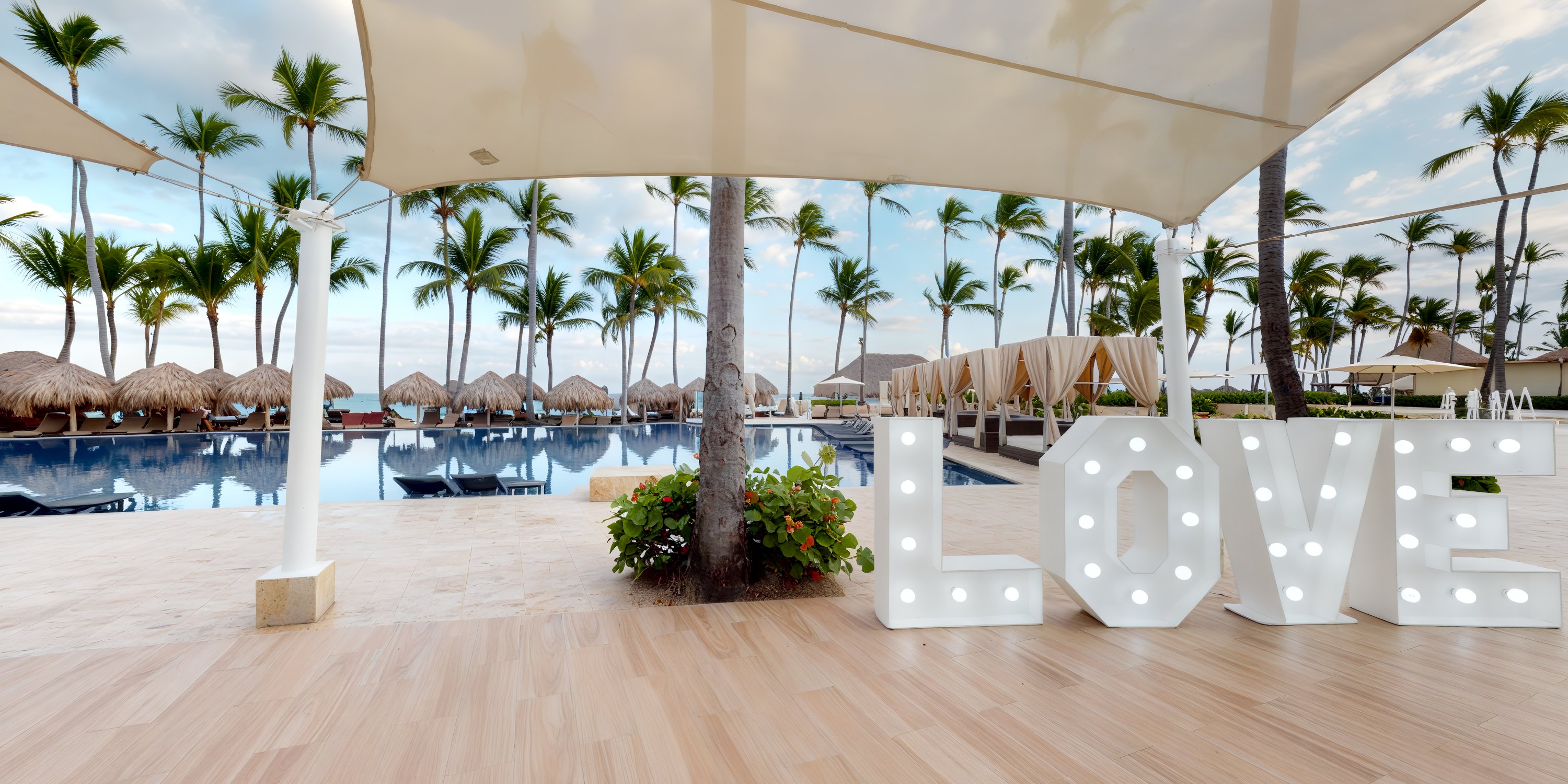 A large "LOVE" sign graces the tropical poolside, surrounded by palm trees and cabanas, creating a perfect setting for a destination wedding.