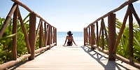 A person stands on a boardwalk gazing at the ocean, donned in swimwear and a UNICO 20°87° hat, perfectly embodying the vibrant spirit of a destination wedding getaway.