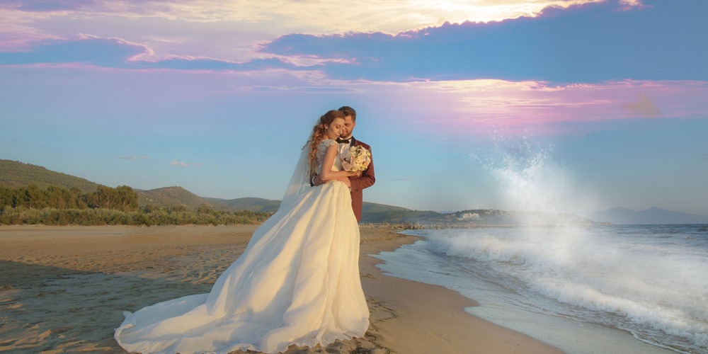 A bride and groom share an embrace on the sandy shore during their destination wedding, with ocean waves and a vibrant sky painting the background.