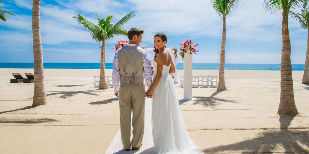 The bride and groom hold hands on a beach surrounded by palm trees and elegant wedding decorations, creating a picture-perfect destination wedding scene.