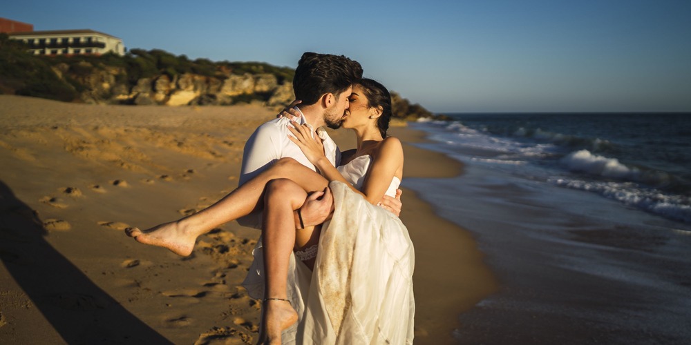 During a romantic destination wedding at sunset, a groom carries and kisses his bride on the sandy beach beside the ocean.