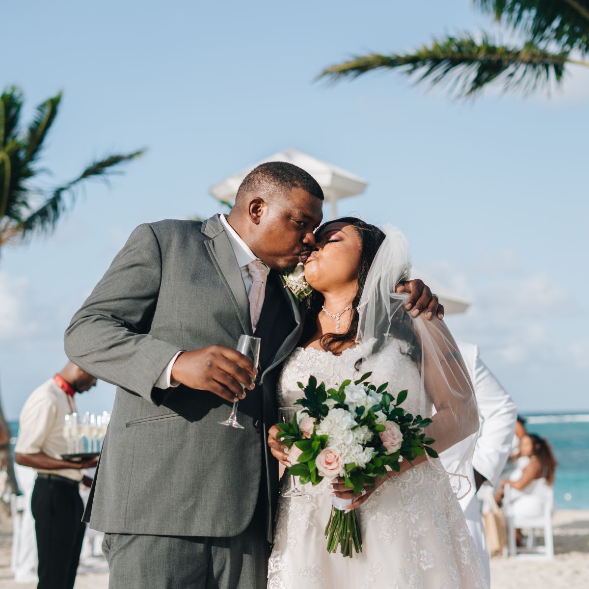 Cheryl and Daryl share a romantic kiss outdoors at their destination wedding, holding a bouquet and a champagne glass.