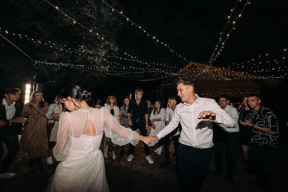 A bride and groom share a romantic dance outdoors at night during their destination wedding, surrounded by guests beneath glowing string lights.