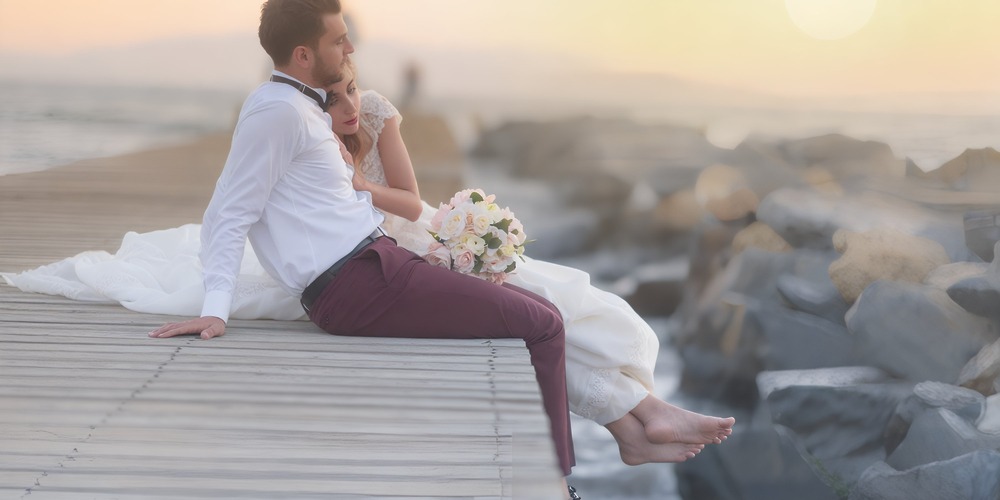 The bride and groom sit together on a pier at sunset, dreaming about a beautiful yet budget-friendly destination wedding.