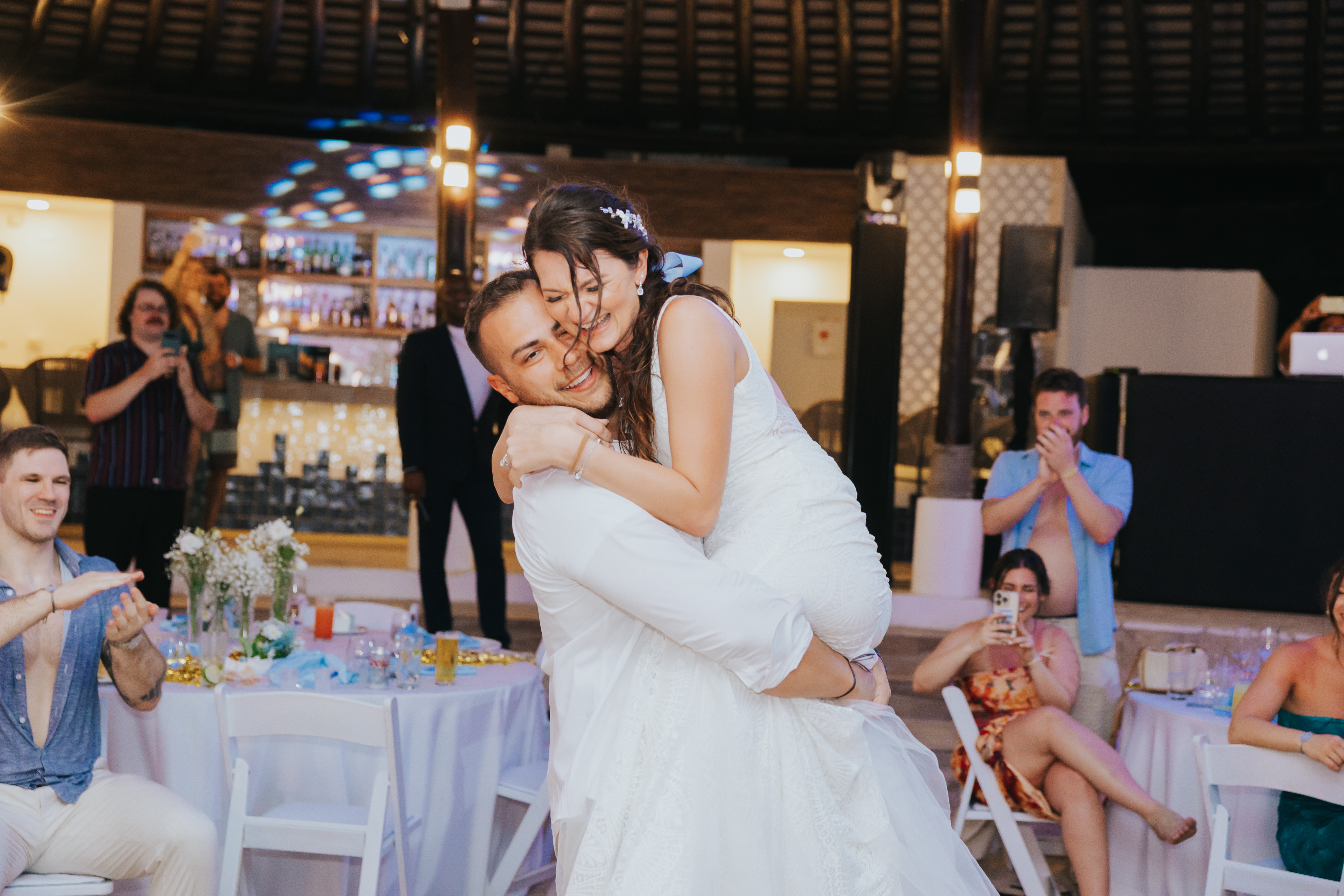 Certainly! Here’s a rewritten version incorporating "wedding" and an optional version with "destination wedding": With “wedding”: At the wedding reception, a groom lifts his bride while guests applaud and capture the joyful moment in photos. With “destination wedding”: During the destination wedding celebration, the groom sweeps his bride into his arms as guests cheer and snap pictures.