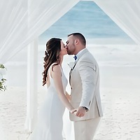 Carina and Steven share a kiss under a white canopy on the beach, holding hands during their destination wedding.