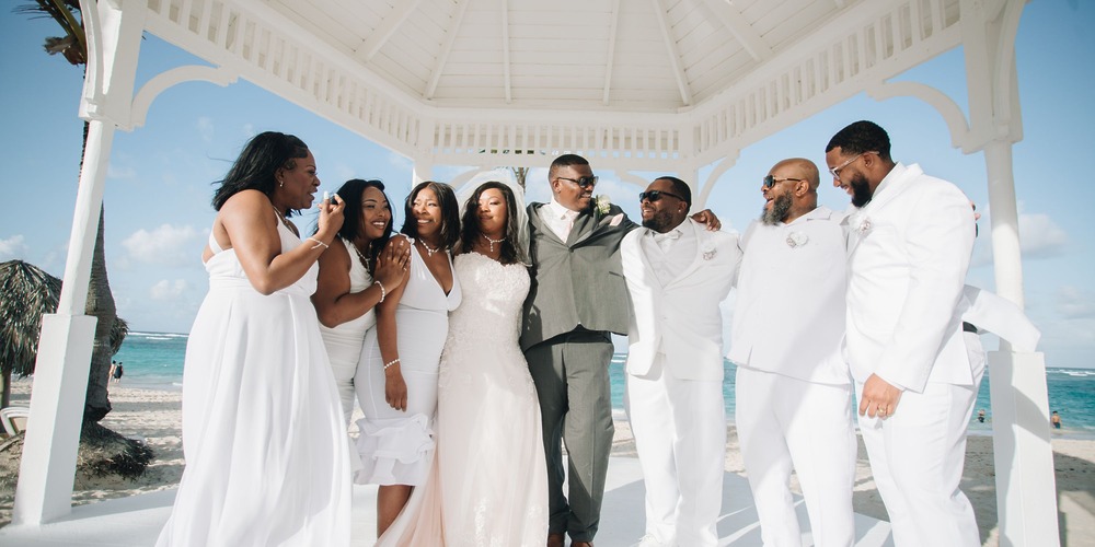 The wedding party gathers beneath a white gazebo by the beach, capturing memories from Cathy and Berwick's beautiful destination wedding.