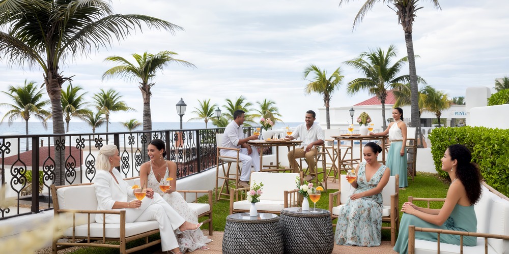 Guests in elegant attire enjoy drinks by the seaside, creating a charming setting that's perfect for a budget-friendly destination wedding.